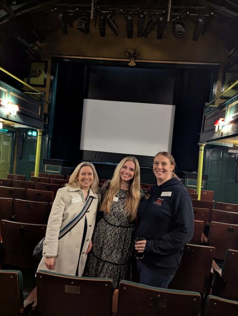 Three women smiling inside a theatre auditorium with a projection screen behind them